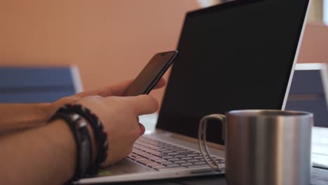 primer plano de manos escribiendo en el teléfono móvil con una taza de acero y una computadora portátil en el fondo