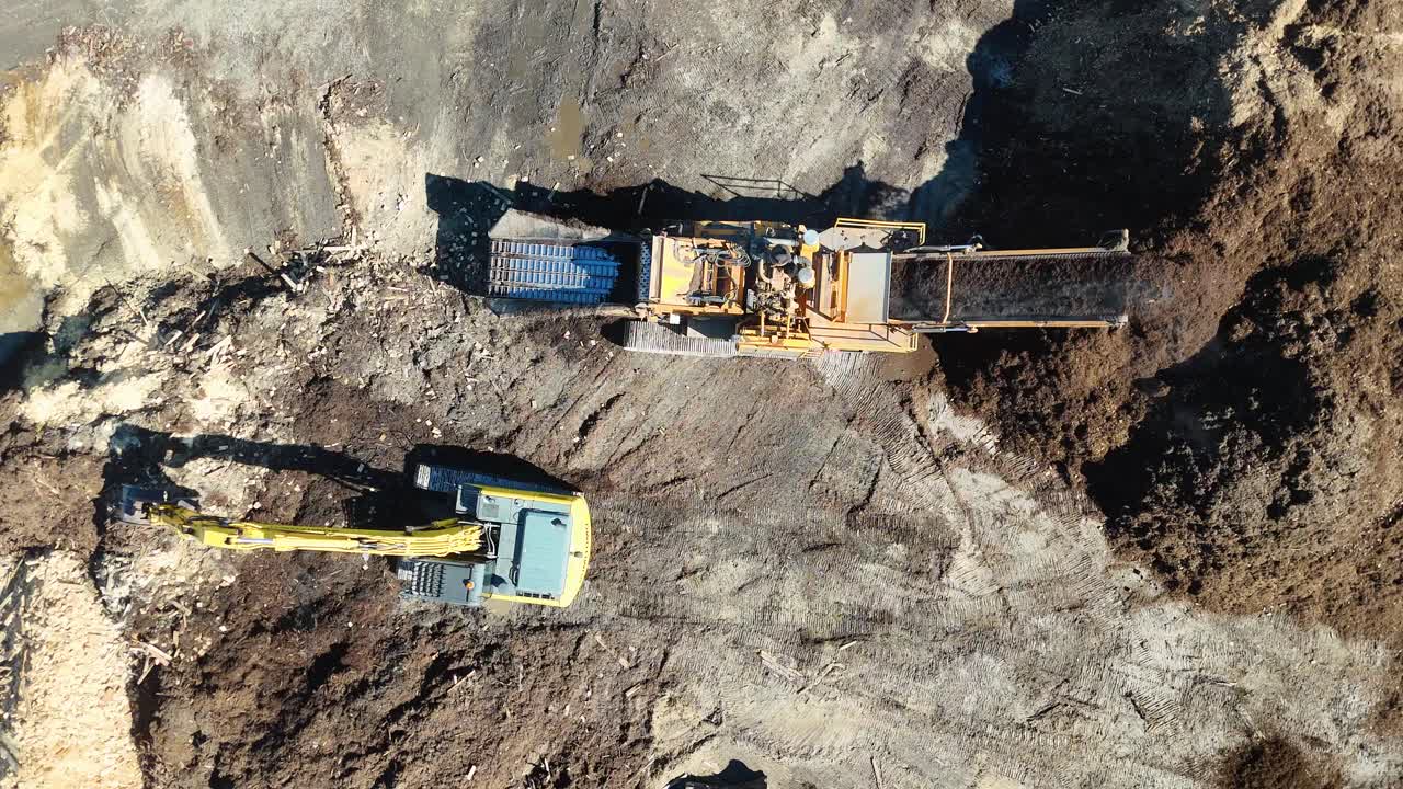 Drone footage captures an excavator moving mulch in a sugar cane field. Bright daylight highlights the machinery and soil