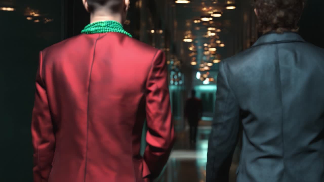 Two individuals in stylish suits walking through a modern hallway adorned with bright lights, showcasing an elegant and fashionable atmosphere