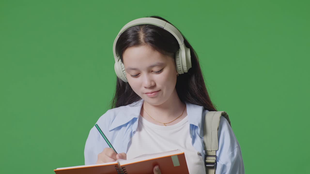 close up de una estudiante adolescente asiática con una mochila con auriculares y tomando notas en el cuaderno mientras está de pie en el estudio de fondo de la pantalla verde