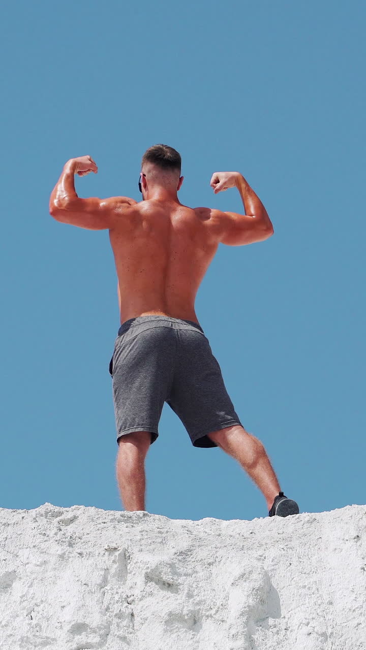 Bodybuilder athlete posing with muscles in nature against a blue sky. A man stands with his back. Vertical video