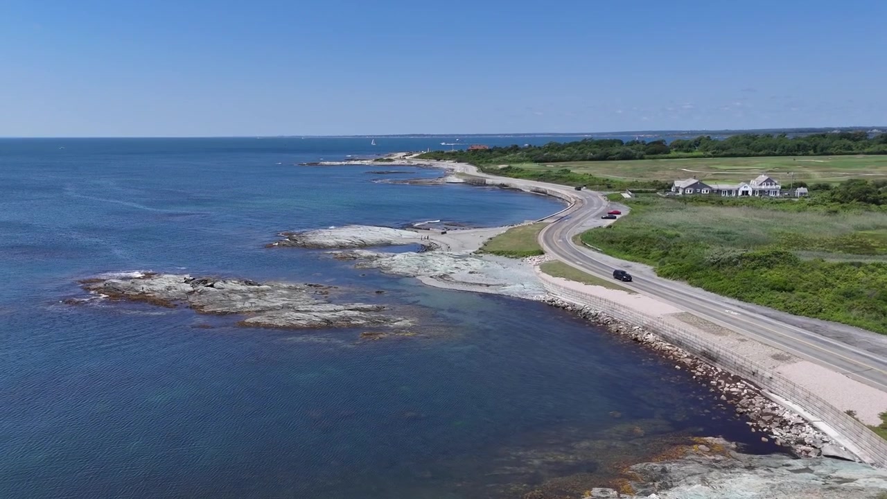Newport, Rhode Island'ın engebeli kayalık kıyı şeridi boyunca lüks deniz kenarı malikanelerini, yeşil çimleri ve manzaralı sahil yolunu görüntüleyen havadan drone takip çekimi