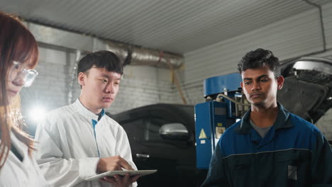 lab technicians observe engineer demonstrating spanner use on car engine, nodding in agreement in mechanical workshop, industrial workspace features vehicles, tools, and equipment in background