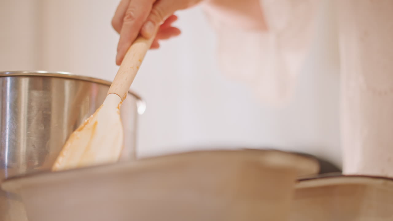 Spatola che livella l'impasto di zucca nelle teglie, ciotola di miscelazione in metallo sullo sfondo, delicato movimento del polso, levigatura finale della superficie, mani infarinate, prontezza per il forno, primo piano caldo