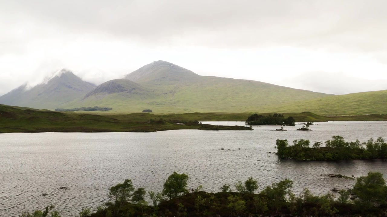 Dron lento se desplaza sobre el lago Rannoch Moor con un pico envuelto en nubes al fondo.