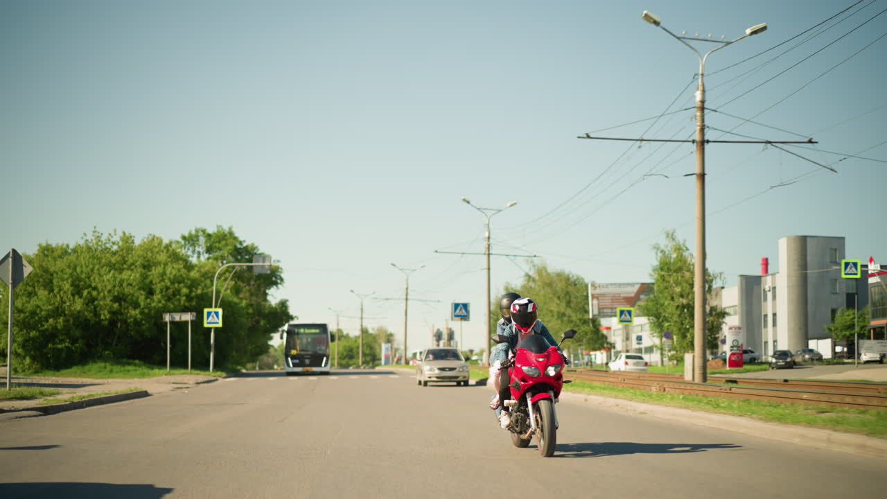 두 친구, 둘 다 헬을 입고, 도시 도로를 따라 빨간색 전력 자전거를 함께 타고, 차와 버스가 뒤에서 따라오고, 도로 건너편에 표지판과 건물이 보입니다.