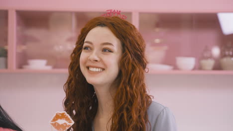 close up view of redhead woman with headdresses and holding cardboard decoration dancing in a pink room celebrating a bachelorette party