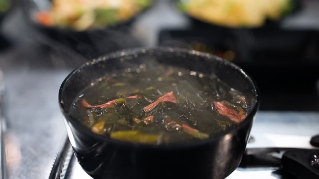 A close-up shot captures steam rising from a small black bowl of traditional Korean seaweed soup called Miyeok Guk served as a side dish at a warm restaurant