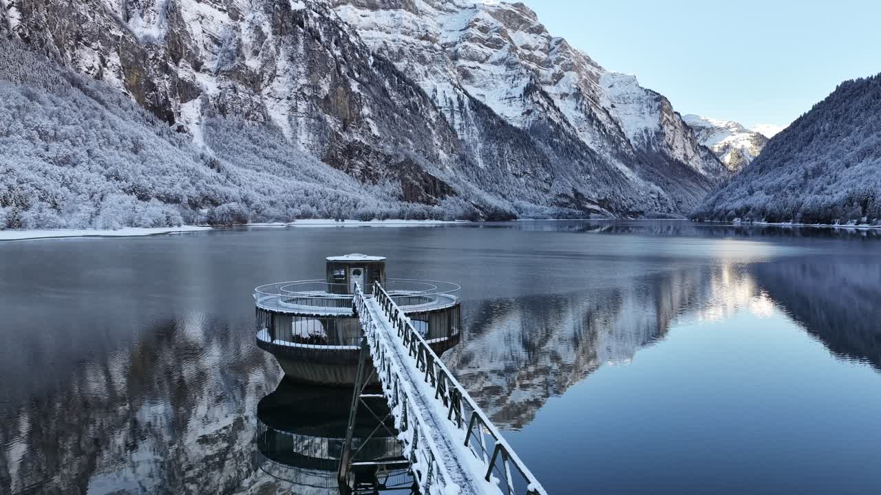 Aerial view of Klöntalersee in Glarus, Switzerland, showing a modern hydroelectric pump house on the dark, reflective lake, framed by snow-covered mountains that merge alpine nature with engineering