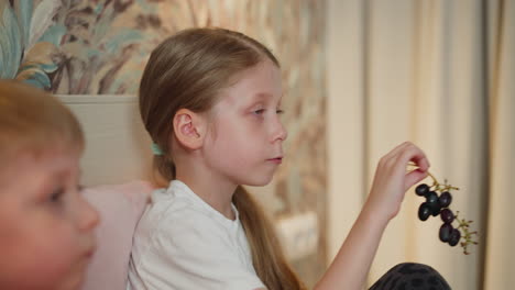 close up of young girl holding black currant branch while picking and eating berries with soft focused sibling beside her sitting on bed in cozy home setting with soft curtain backdrop