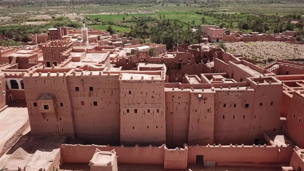 Vista drone di una grande fortezza kasbah circondata da una lussureggiante vegetazione di oasi e paesaggio vallivo a Ouarzazate, Marocco. Forte contrasto tra architettura storica e ambiente naturale, ideale per
