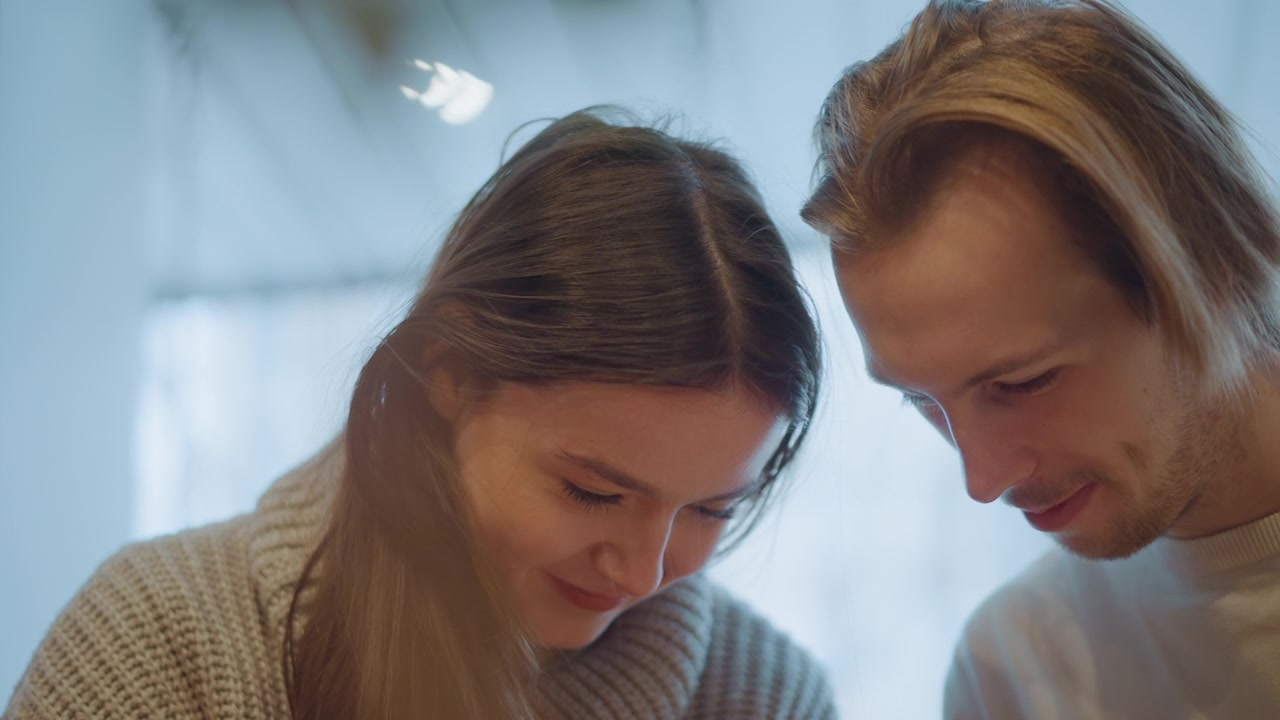 Primo piano di due persone, una donna e un uomo con lunghi capelli, che guardano in basso insieme in una morbida illuminazione interna. La scena li mostra vicini, fianco a fianco, contro uno sfondo sfocato con luce diffusa.