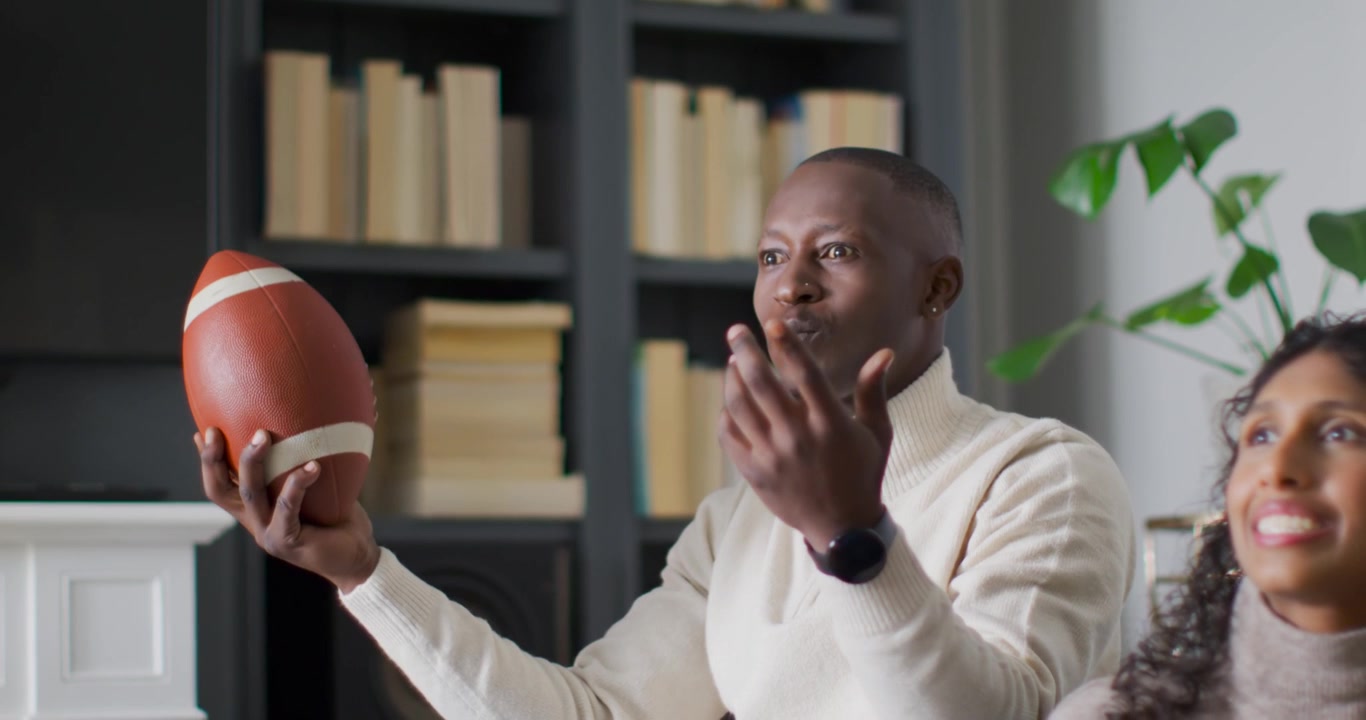 African American couple reacting to on-screen game in living room, man holding football, gesturing