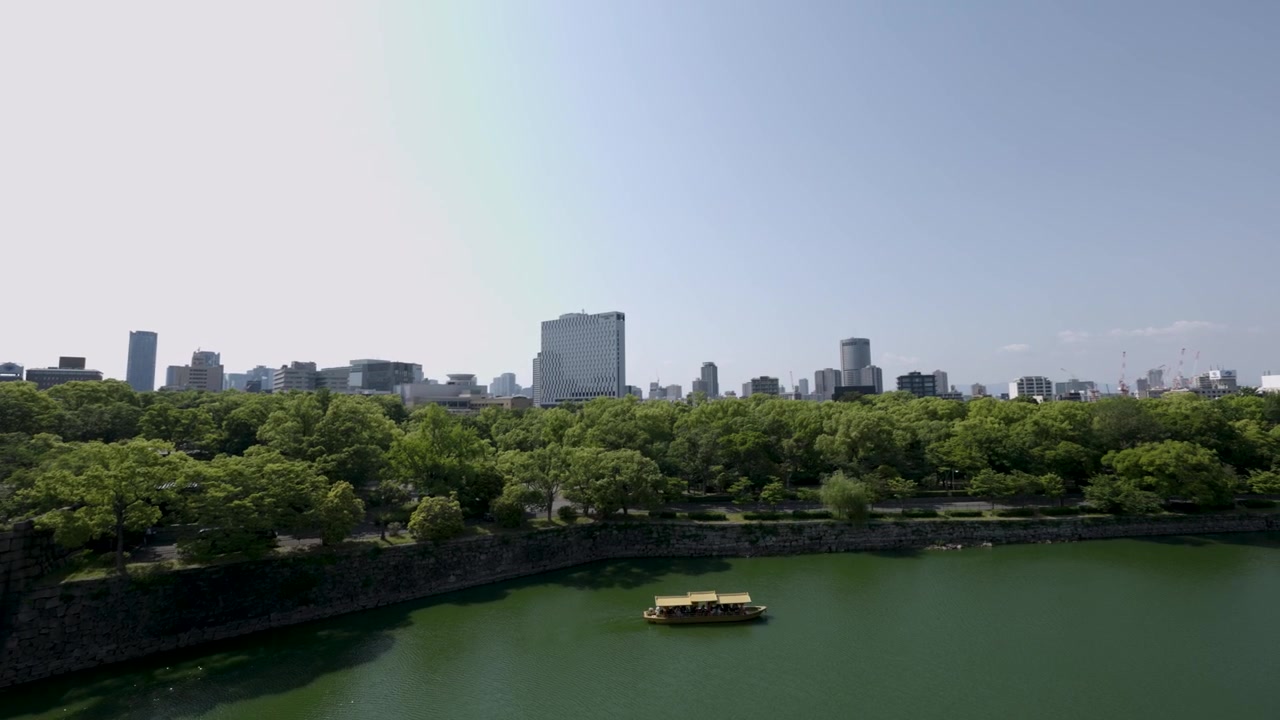 Osaka Kalesi'nin hendeklerinde bir tekne, yeşillikler ve Osaka şehir manzarası.