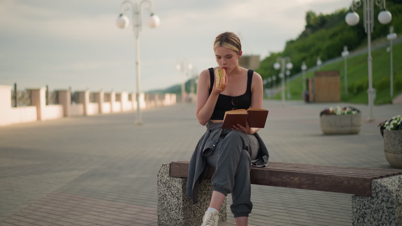 женщина сидит на открытом воздухе с перекрестными ногами, держит книгу в одной руке, в то время как ест закуски и читает, размытый фон включает в себя фонарь и зеленый холм