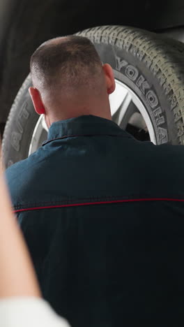vista de fondo de un inventor en bata de laboratorio fotografiando a un mecánico arreglando un neumático de coche con un teléfono inteligente mientras un estudiante mira, con los brazos cruzados, en un taller mecánico