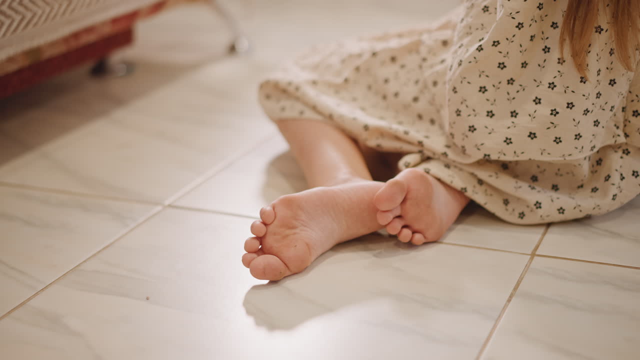Bare feet child sitting on tile, soft sunlight across patterned dress, relaxed posture near radiator, closeup of toes and floor texture, warm cozy domestic mood suggesting quiet daydream