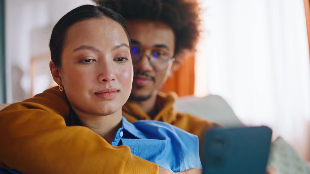 Mixed race couple browsing smartphone on sofa at home. Closeup happy pair.