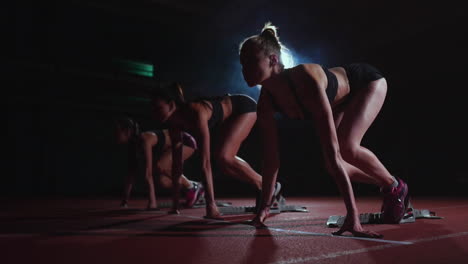 Three-girls-in-black-clothes-are-in-the-starting-pads-to-start-the-race-in-the-competition-in-the-light-of-the-lights-and-run-towards-the-finish