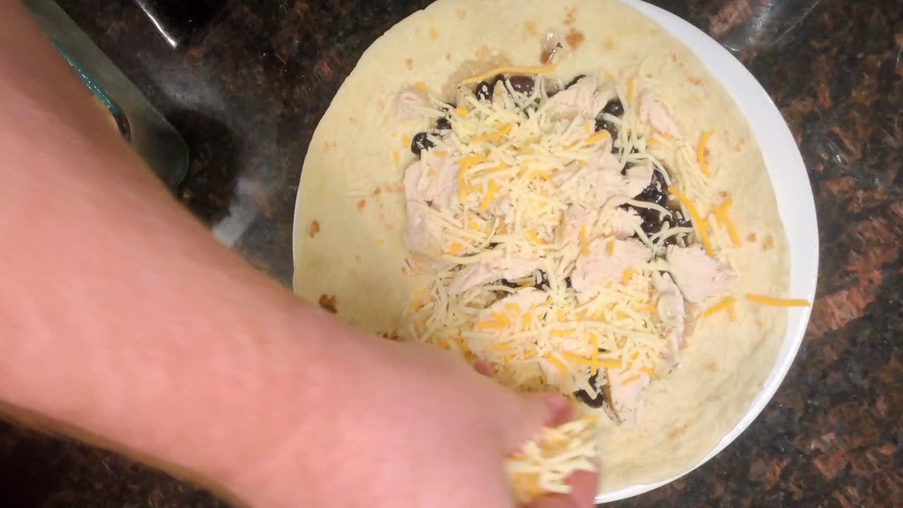 Close-up top-down footage captures a chef’s hands sprinkling cheese onto a tortilla wrap in a kitchen