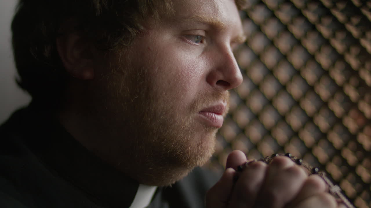 Close Up of Priest Holding Rosary while Praying inside Confessional Booth