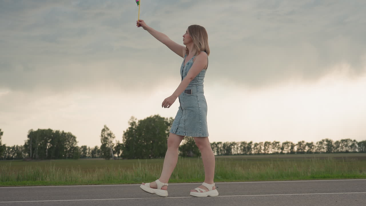 Side view of woman walking along white road marking holding colourful pinwheel rotating under cloudy summer sky beside open grassy field with distant trees hair swaying in gentle breeze