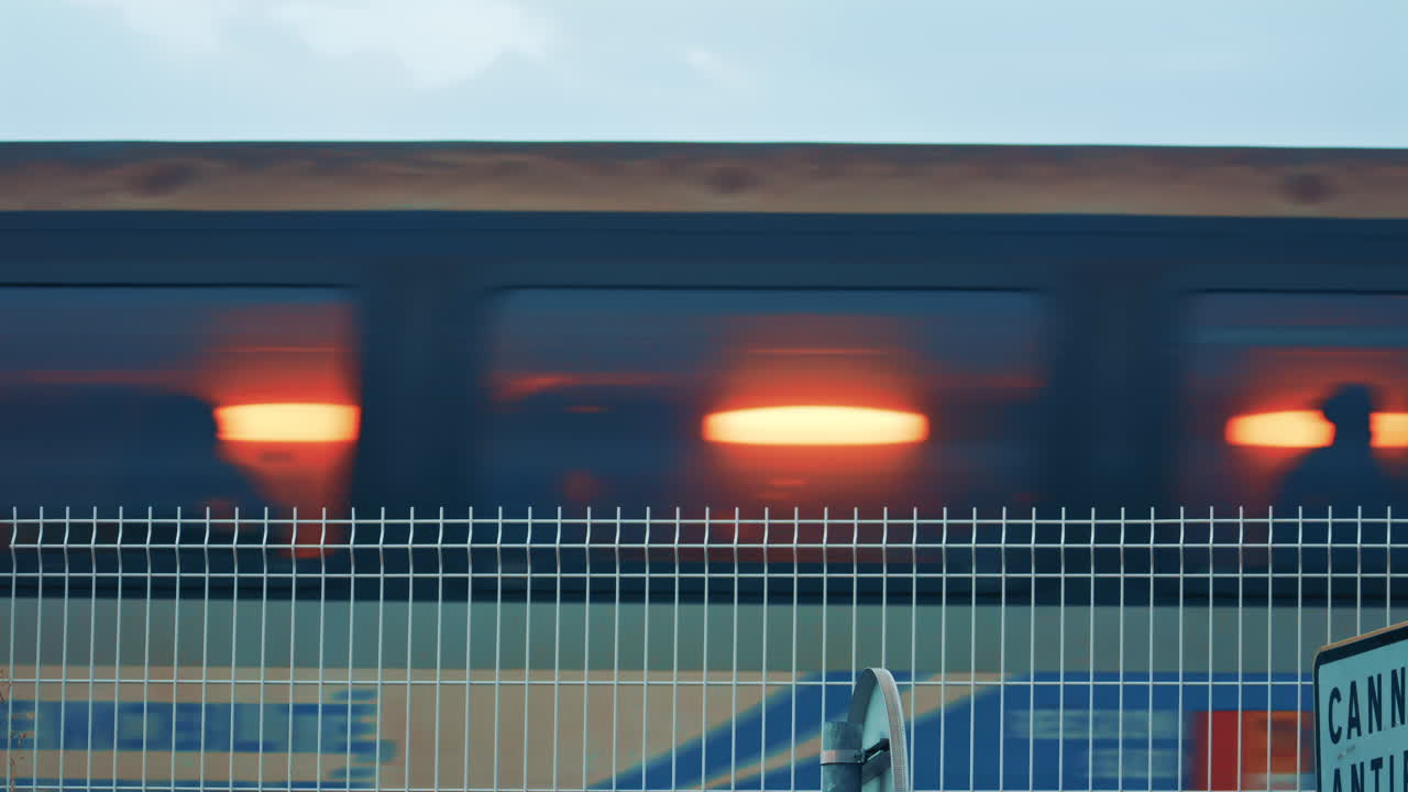 A fast passenger train rushing past behind a metal fence, with blurred motion and glowing interior light