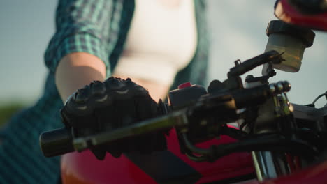 close-up of a woman in a green checkered shirt, white crop top, and protective gloves throttling a red motorcycle