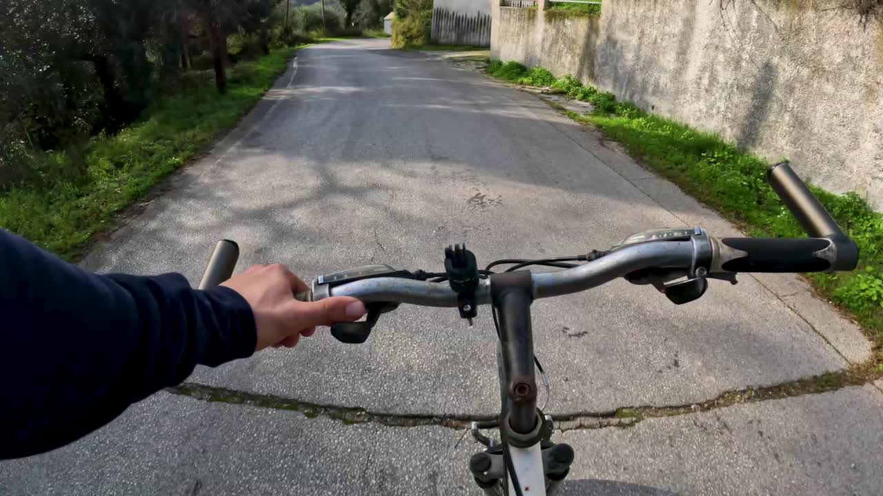 Ciclismo en primera persona cuesta arriba en carretera de asfalto desgastado en zona rural verde, ciclista POV montando en bicicleta pasando muros de piedra y árboles, entrenamiento físico al aire libre en la naturaleza Corfú Grecia