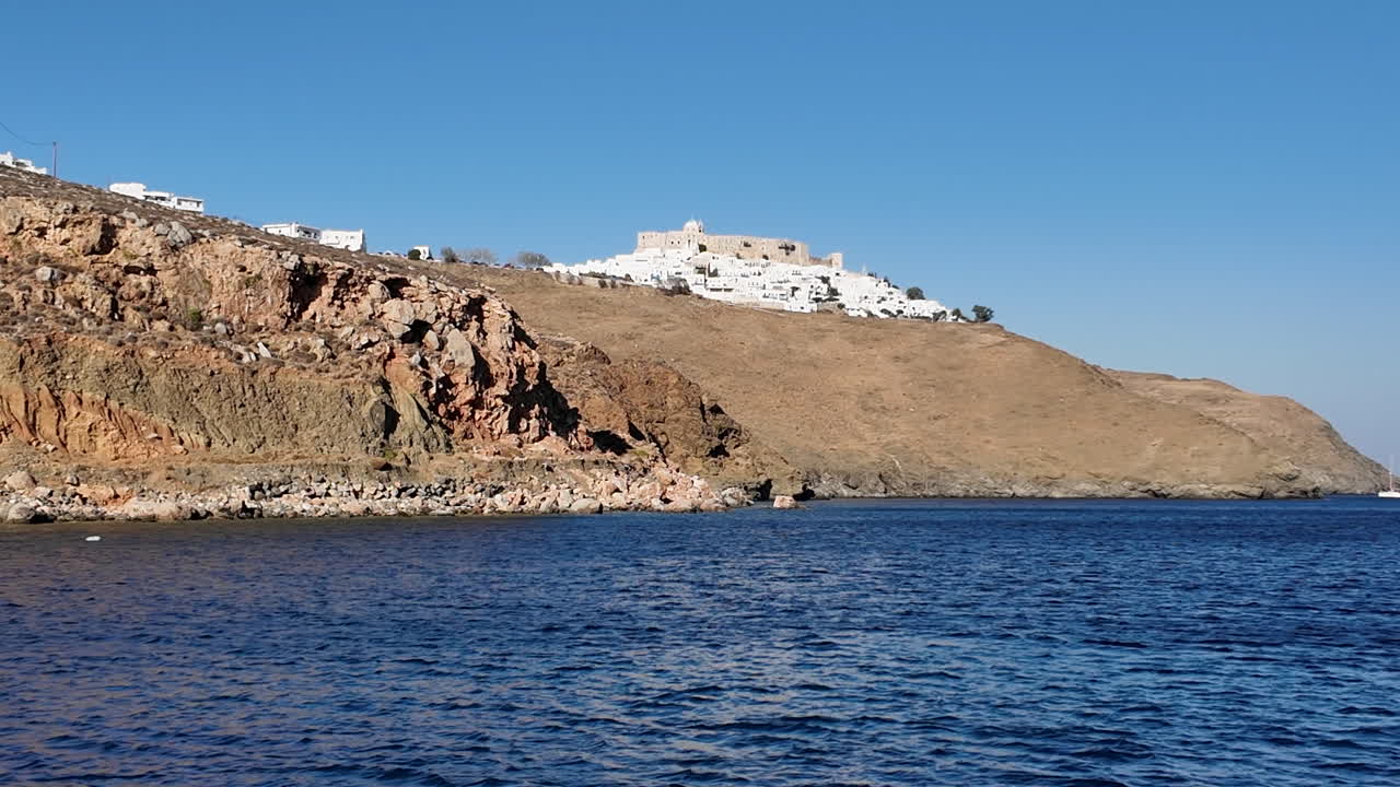 Greece, Astypalea Island, Livadi Bay, from the sea, beach, trees, boats and flight passing over the iconic Castle on a sunny day with blue skies.