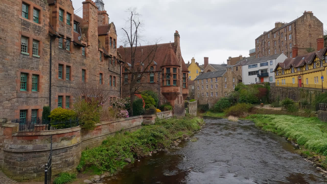Slow motion pan across flowing river bubbling on mossy shores between tall old brown brick buildings of Edinburgh Scotland