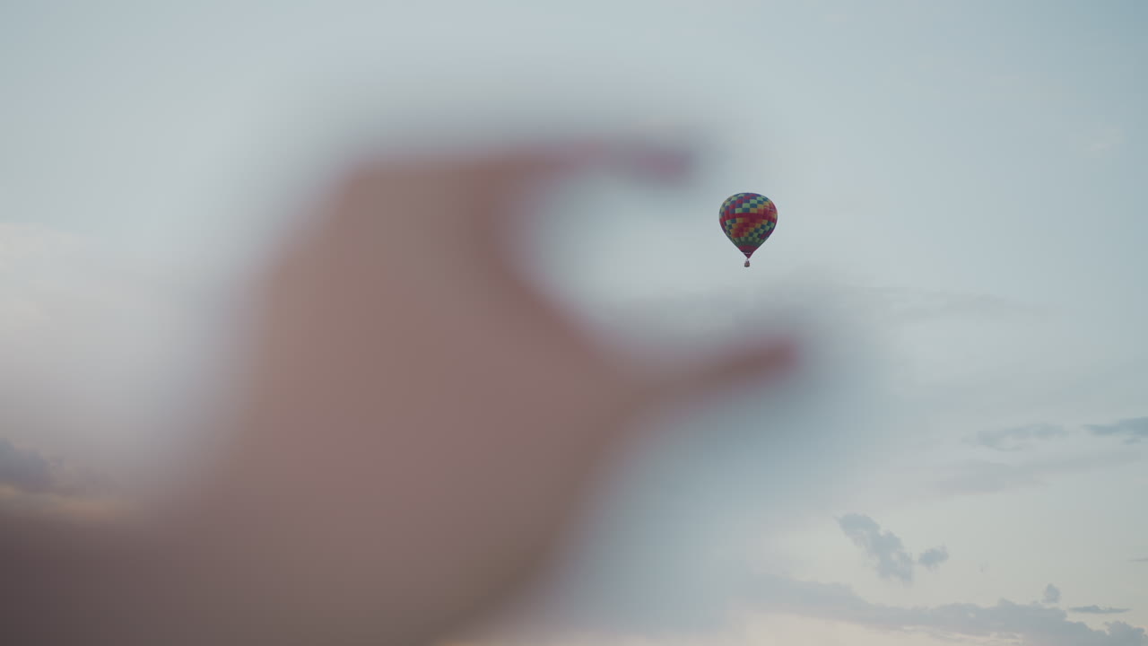 blurred hand entering frame reaching toward colorful hot air balloon drifting across soft pastel twilight sky over tranquil horizon with distant clouds evoking wonder and travel perspective