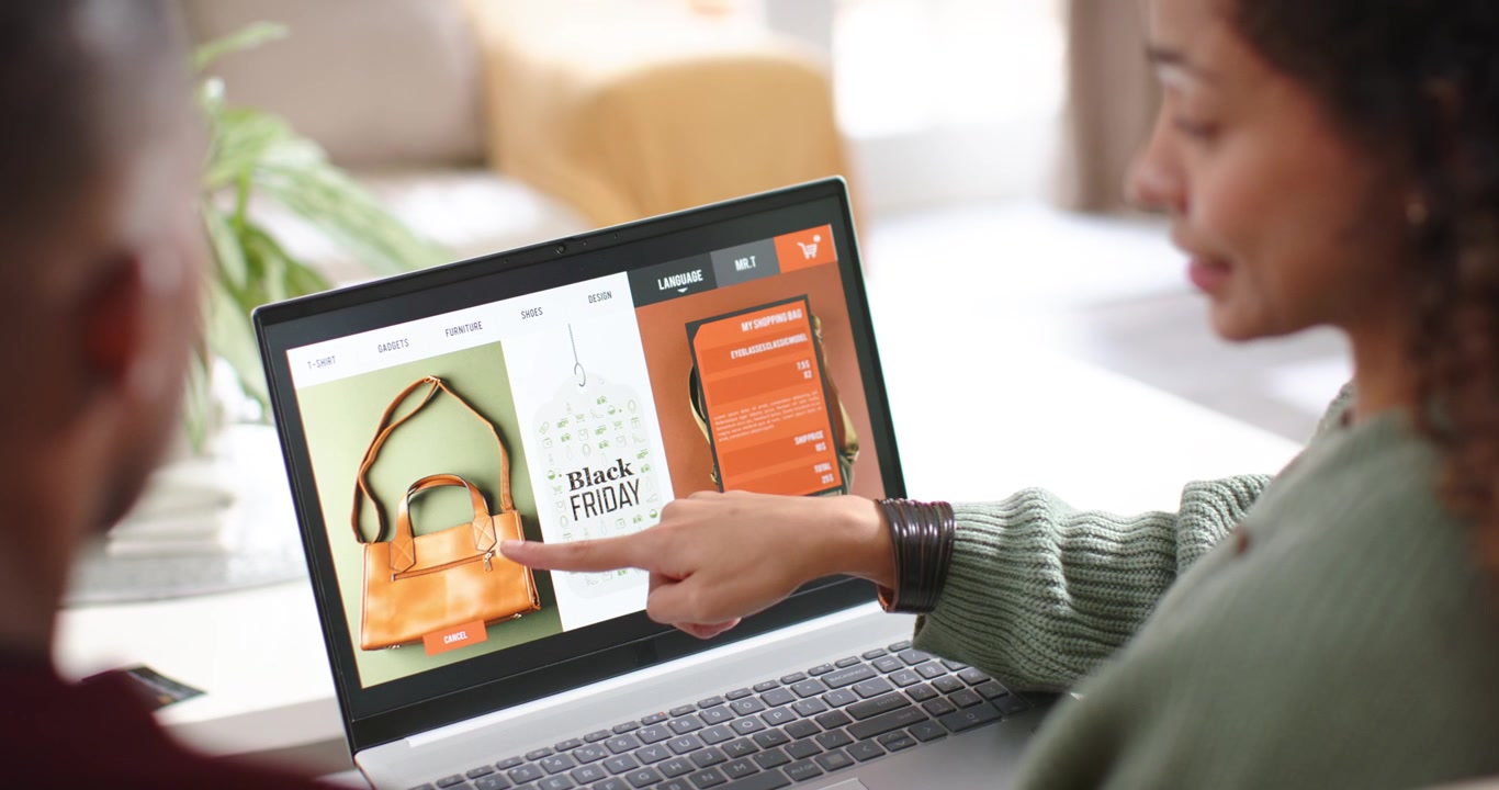 Coppia che naviga per il Black Friday al laptop a tavola, la donna che indica, toccando il pulsante "aggiungi al carrello" per una borsa per acquistarla