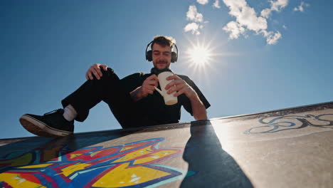 young man relaxing with headphones and a mug at a sunny skatepark