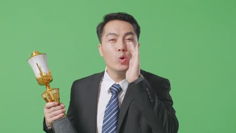 슈트와 타이를 입은 아시아 사업가, 금메달을 들고, 금 트로피를 들고, 녹색 스크린 배경에서 입을 고 긍정적으로 소리치고 있습니다.