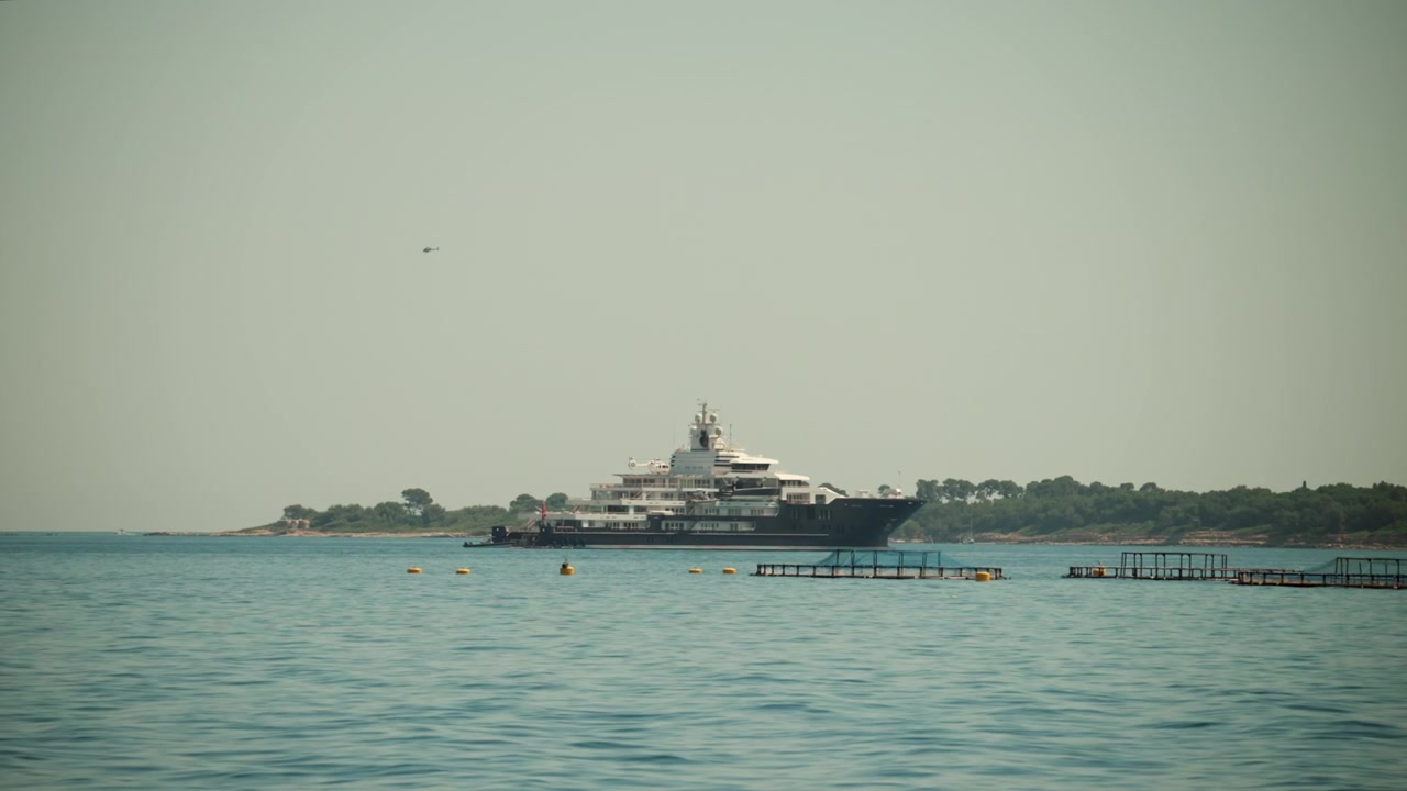 Super yacht ormeggiato in calme acque blu del Mar Mediterraneo, che mostra viaggi di lusso e ricchezza in contrasto con le gabbie per l'allevamento di pesci