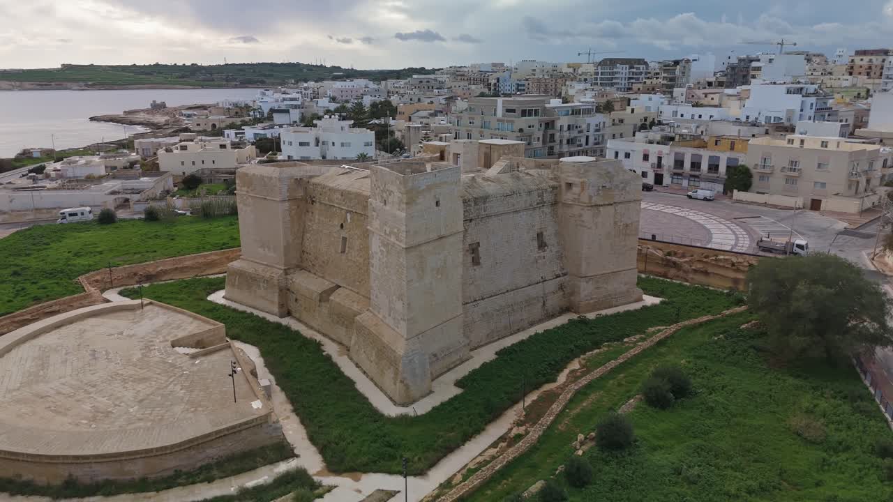 Vista de dron en órbita de Torri San Tumas, una torre Wignacourt del siglo XVII en Marsaskala, Malta, que muestra su arquitectura histórica de piedra caliza en medio de un paisaje costero y urbano mediterráneo