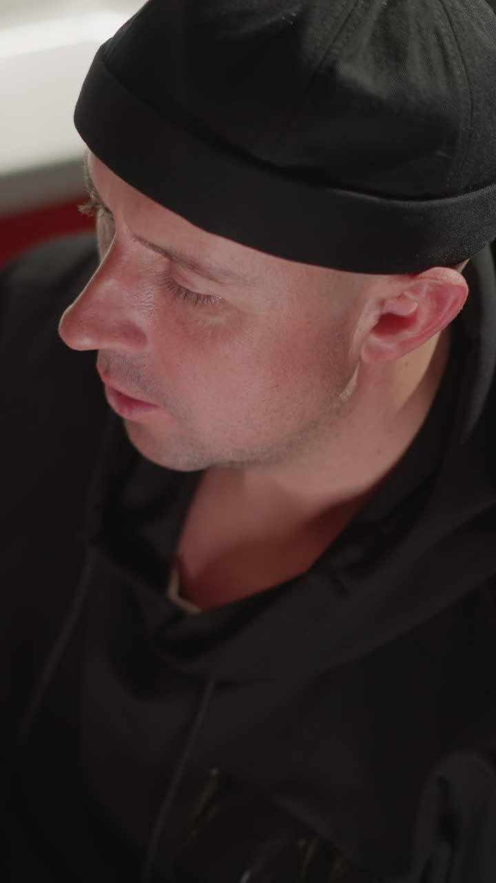 A man dressed in black is sitting at a desk in a recording studio, focused on working at a computer. The setting includes sound equipment, with another person blurred in the background