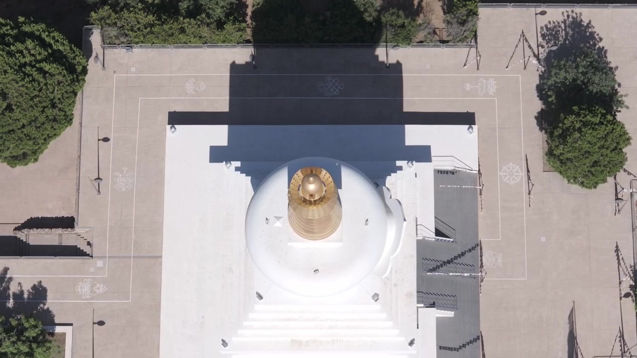 Images de drone plongeant sur la flèche annelée d'or et le dôme blanc de la Stupa de Benalmádena, révélant la cour intérieure, la garrigue sèche et les arbres environnants en contrebas.
