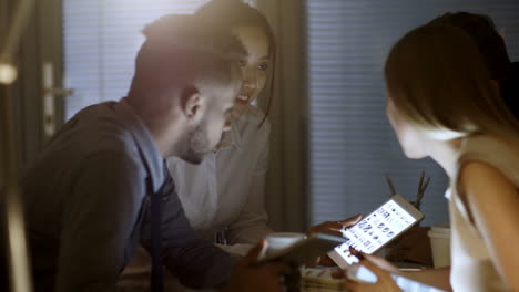 young woman showing photos on tablet computer to group of multi ethnic friends or colleagues late in the evening