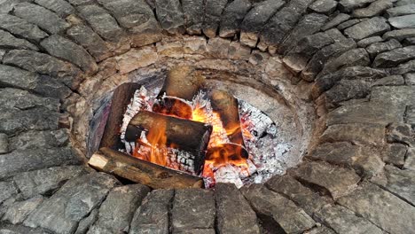 Beautiful-circular-stone-fire-pit-with-flames,-hot-coals-and-burning-logs-during-a-cold-Winters-day-outdoors-in-the-garden