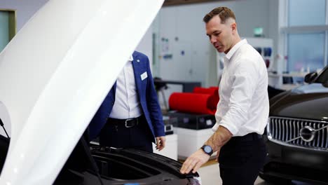 salesman showing car engine under open hood to a client in auto showroom