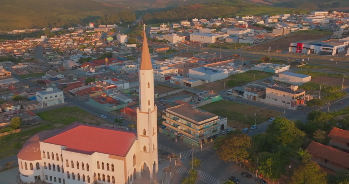 Gereja Santo Antonius menghadap jalan-jalan pusat kota dan blok perumahan di Santo Antônio do Monte, Minas Gerais, Brasil, mendokumentasikan arsitektur perkotaan saat cahaya matahari terbenam yang hangat, dengan pendakian mundur drone.