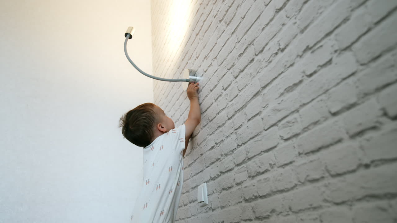 Happy active baby boy jumps excitedly on the bed. Kid plays with the lamp turning on and off the light. Low angle view.