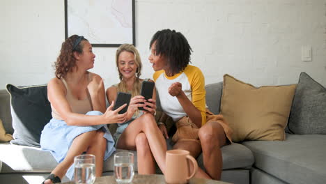 diverse female friends laughing on sofa, sharing moments on smartphones, friendship, at home