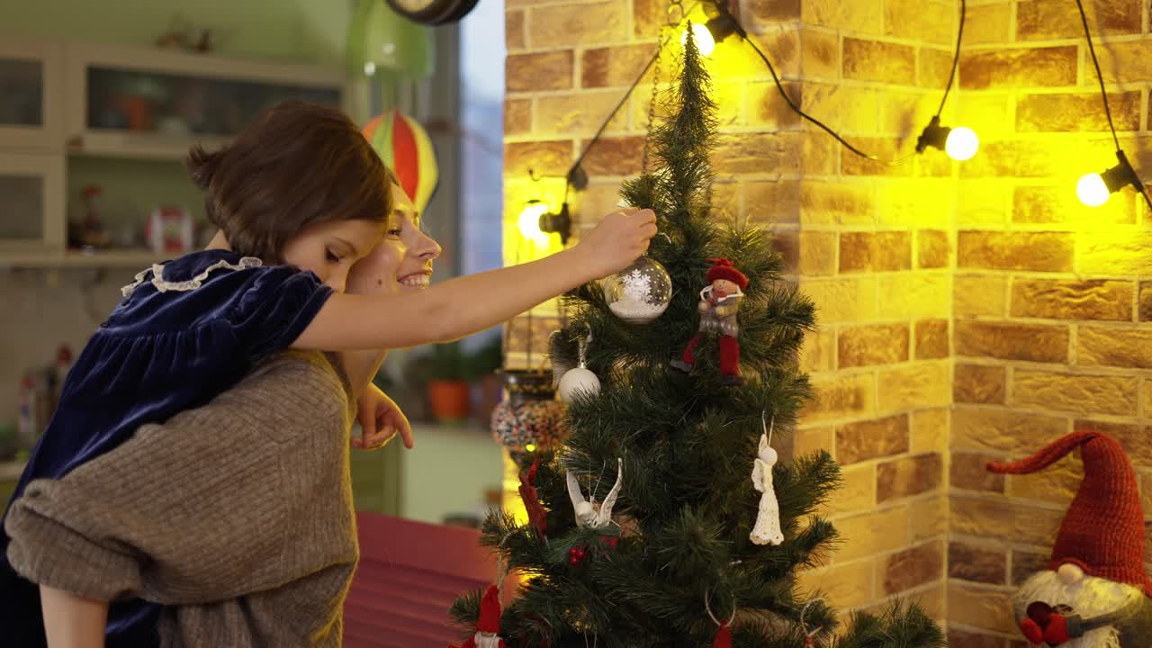 madre sosteniendo a la niña en la espalda, colgando juntos los juguetes del árbol de navidad encima del árbol de navidad.