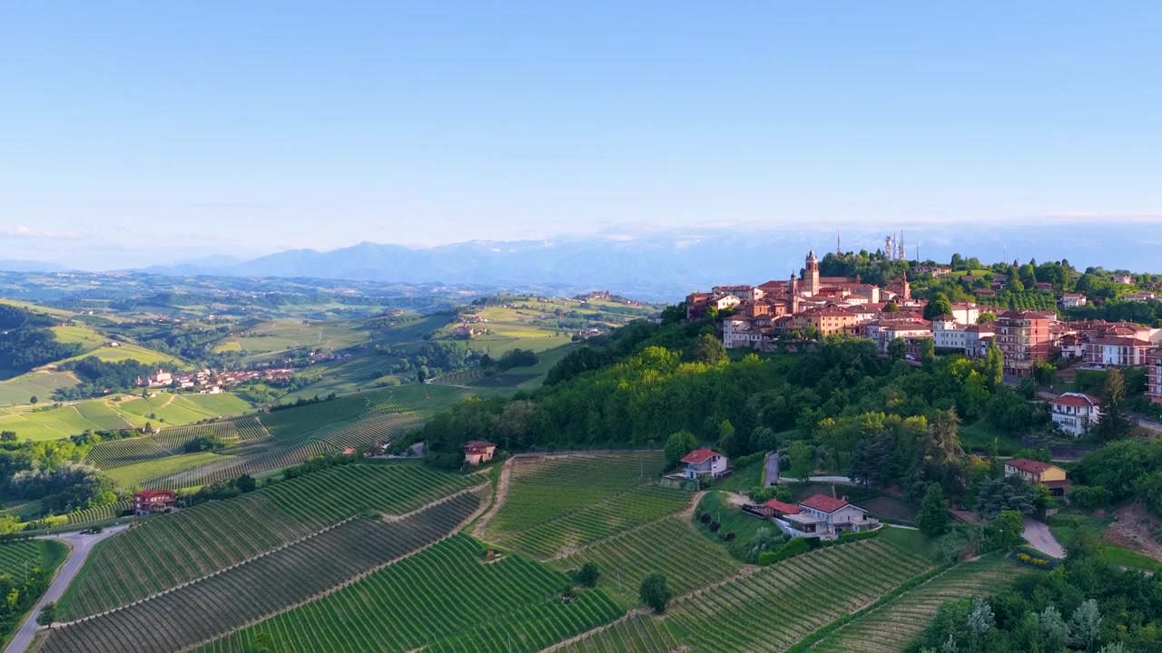 Ripresa aerea con drone di un borgo storico in cima a una collina che domina rigogliosi vigneti verdi in Piemonte, Italia.
