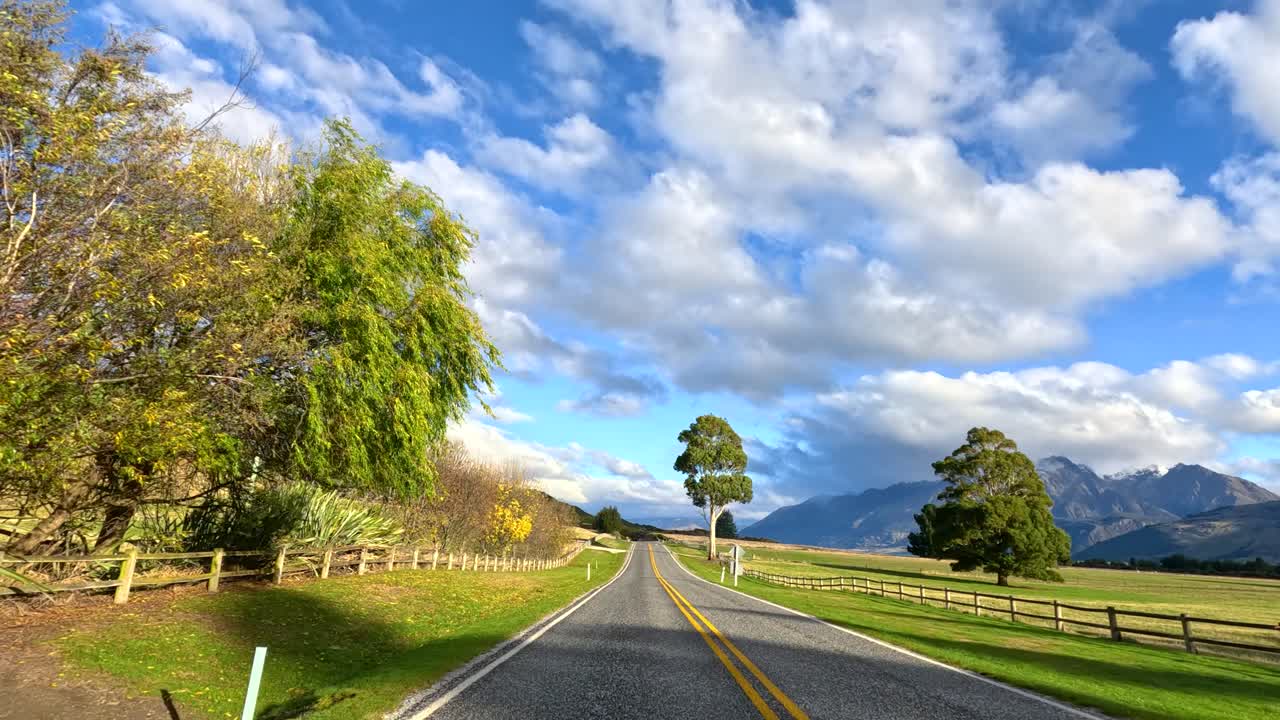 Vehicle travels rural road with mountain views, green fields, wooden fences, and bright daylight