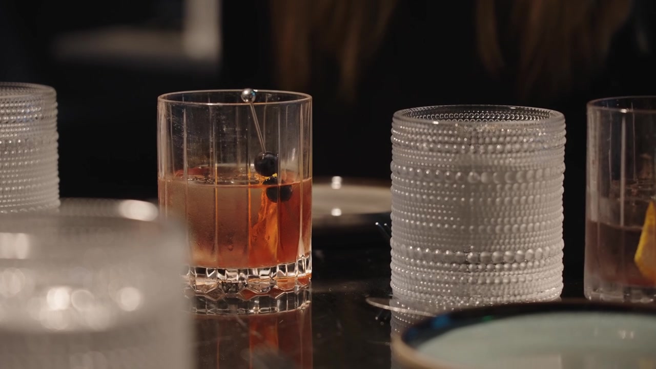 Transizione di messa a fuoco 4K di un cocktail Old Fashioned guarnito con mirtilli e scorza d'arancia in un'ambientazione suggestiva di un hotel di lusso, che evidenzia la mixology raffinata e i dettagli cinematografici.