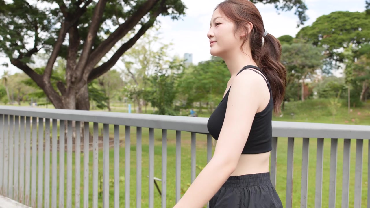 緑豊かな公園で、黒いアクティブウェアを着た若いアジア人女性が手すりのそばをジョギングしている。